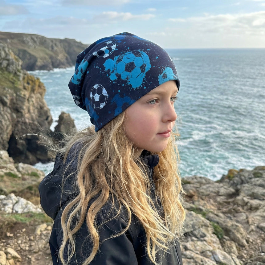 Kind mit blonden Haaren trägt Wollhuhn Öko Long Beanie, zweilagig, mit Fußballmotiven, ideal für das ganze Jahr. Steht am felsigen Küstenmeer.