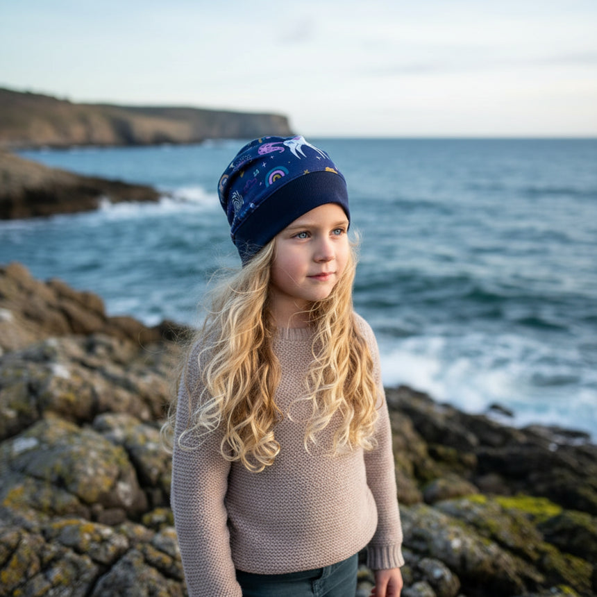 Mädchen mit blonden Haaren trägt eine Wollhuhn Dünne Öko Beanie-Mütze mit Einhornmotiv am Meer, ideal für Übergangszeit, handgefertigt in Deutschland.