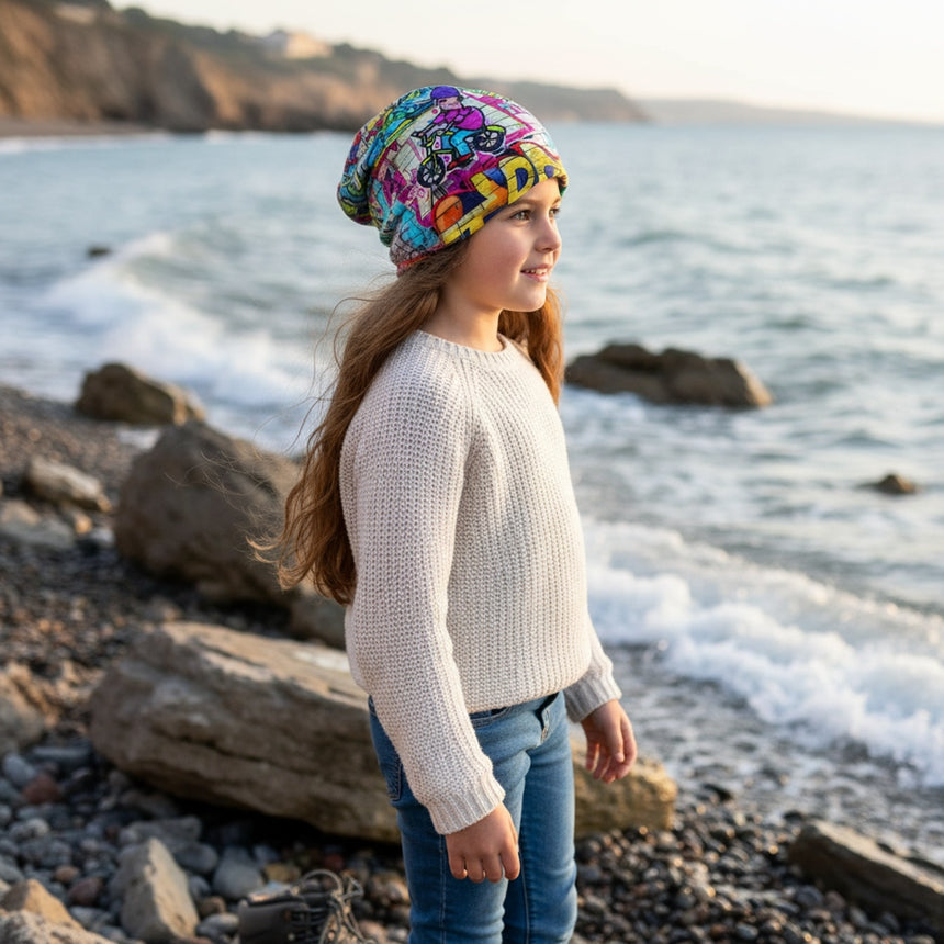Mädchen am Strand trägt die Wollhuhn Öko Long Beanie, eine zweilagige, bunte Wendemütze, ideal für das ganze Jahr, bequem und wachstumsfreundlich.