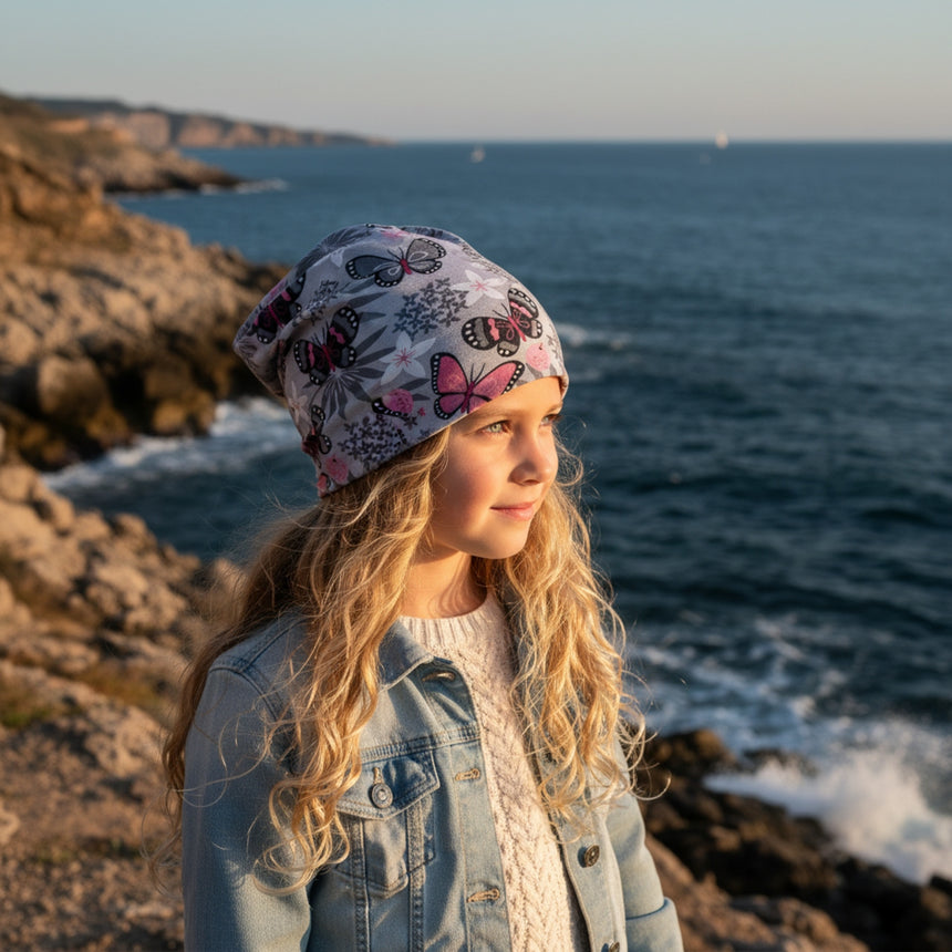 Mädchen am Meer trägt Wollhuhn Öko Long Beanie, zweilagig, schmetterlingsmotiviert, lässig über den Kopf fallend. Perfekt für stilvolle und bequeme Übergangszeiten.