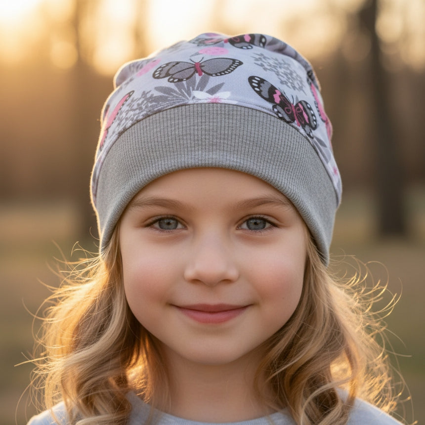 Lächelndes Mädchen trägt Wollhuhn Dünne Öko Beanie-Mütze Sommer/Übergang Einlagig Schmetterlinge Grau/Rosa im Park, handgemacht und nachhaltig, ideal für Übergangszeit.