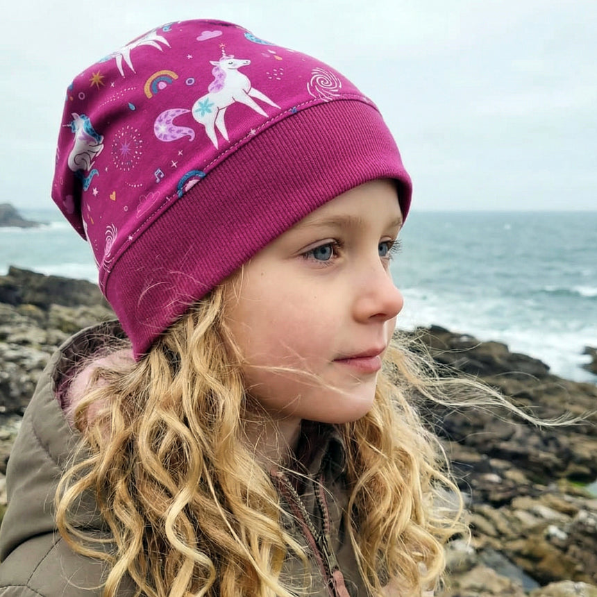 Kind am felsigen Strand mit einer Wollhuhn Dünne Öko Beanie-Mütze, einlagig mit Einhörnern und Regenbögen, ideal für Übergangszeit, handgefertigt in Deutschland.