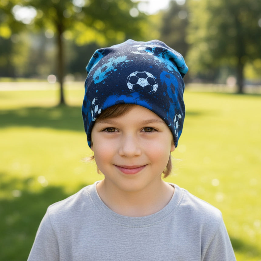 Kind trägt Wollhuhn Öko Long Beanie mit Fußballmuster im Park, lächelnd. Mütze aus weichem, zweilagigem Jersey, angenehm und ganzjährig tragbar.