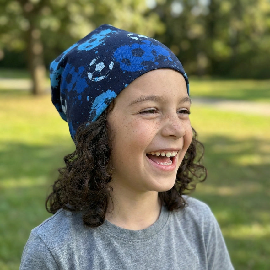 Kind mit Wollhuhn Öko Long Beanie, Wendemütze, Fußball Blautöne, lächelt im Freien. Beanie aus weichem, zweilagigem Jersey, ideal für ganzjährigen Tragekomfort.