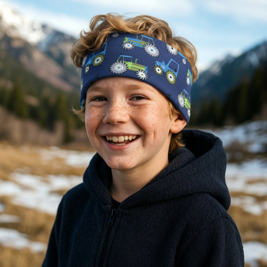 Junger Junge trägt Wollhuhn Elastisches Öko Stirnband mit Traktor-Muster, steht lächelnd in winterlicher Berglandschaft. Stirnband aus weicher, atmungsaktiver Bio-Baumwolle, ideal für Outdoor-Aktivitäten.