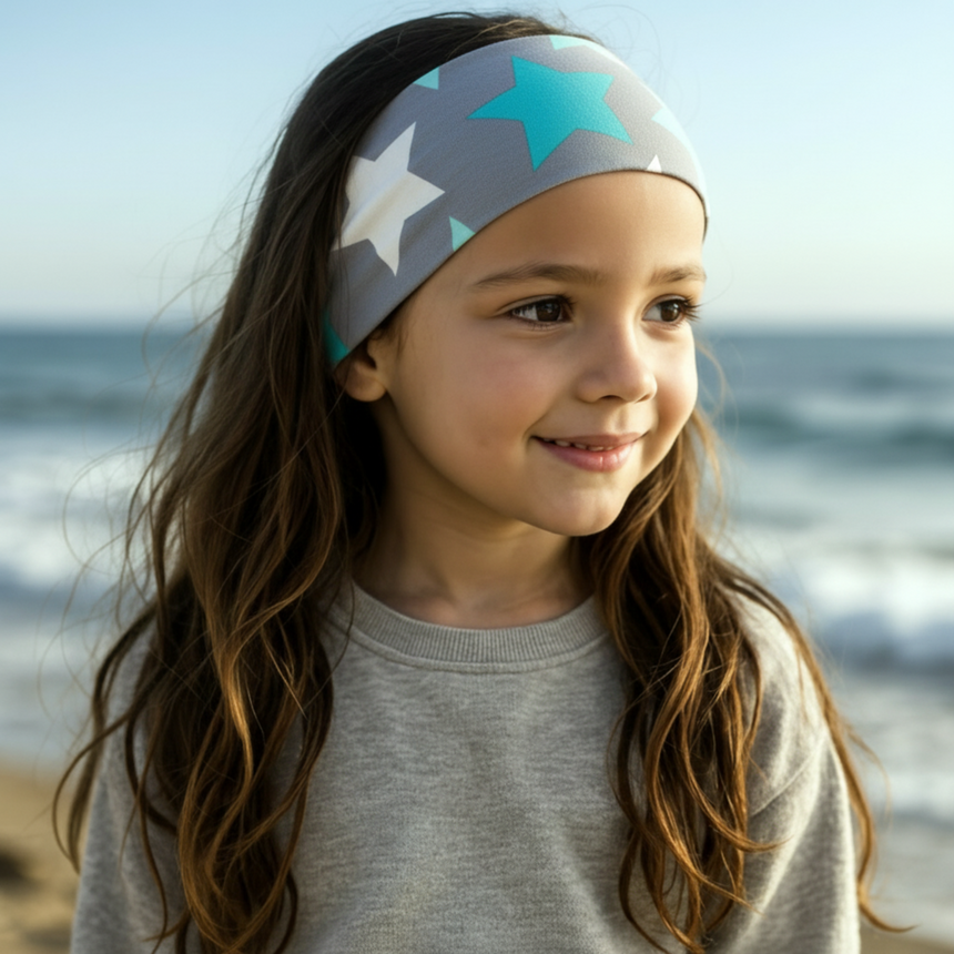 Mädchen am Strand trägt Wollhuhn Elastisches Öko Stirnband/Haarband Jersey Zweilagig Breit Big Stars Sterne Grau/Mint, ideal für Outdoor und Aktivitäten.