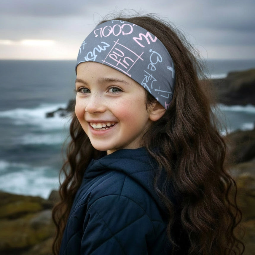 Mädchen am felsigen Ufer trägt Wollhuhn Elastisches Öko Stirnband / Haarband Jersey Zweilagig Breit Cool Music Grau/Rosa, lächelt mit windgekämmtem Haar vor Meerkulisse.