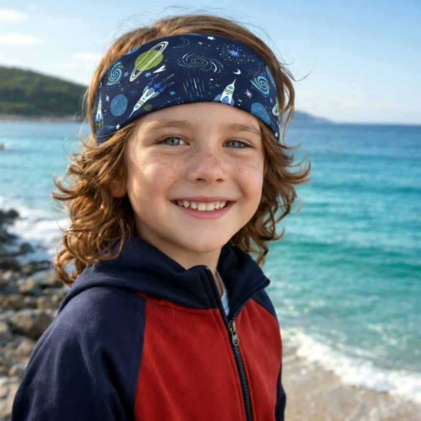 Ein Junge am Strand trägt das Wollhuhn Elastisches Öko Stirnband/Haarband Jersey Zweilagig Breit Space/Rakete Dunkelblau, ideal für Outdoor-Aktivitäten.