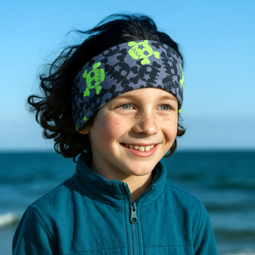 Junge am Wasser trägt Wollhuhn Elastisches Öko Stirnband Jersey Zweilagig Breit mit Totenköpfen, ideal für Outdoor-Aktivitäten, handgemacht aus weicher Bio-Baumwolle in Deutschland.