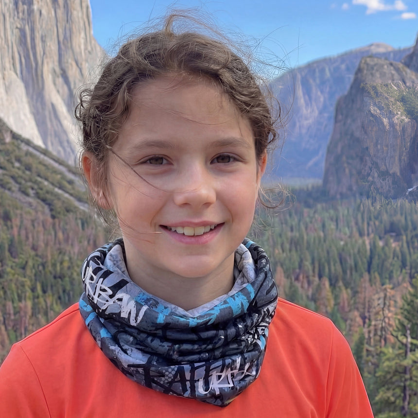 Junges Mädchen trägt Wollhuhn LONG Loop, Jersey- oder Fleecefutter, Skater Blau, in einer Waldlandschaft, lächelt in die Kamera. Perfekt für Kinder und Erwachsene.