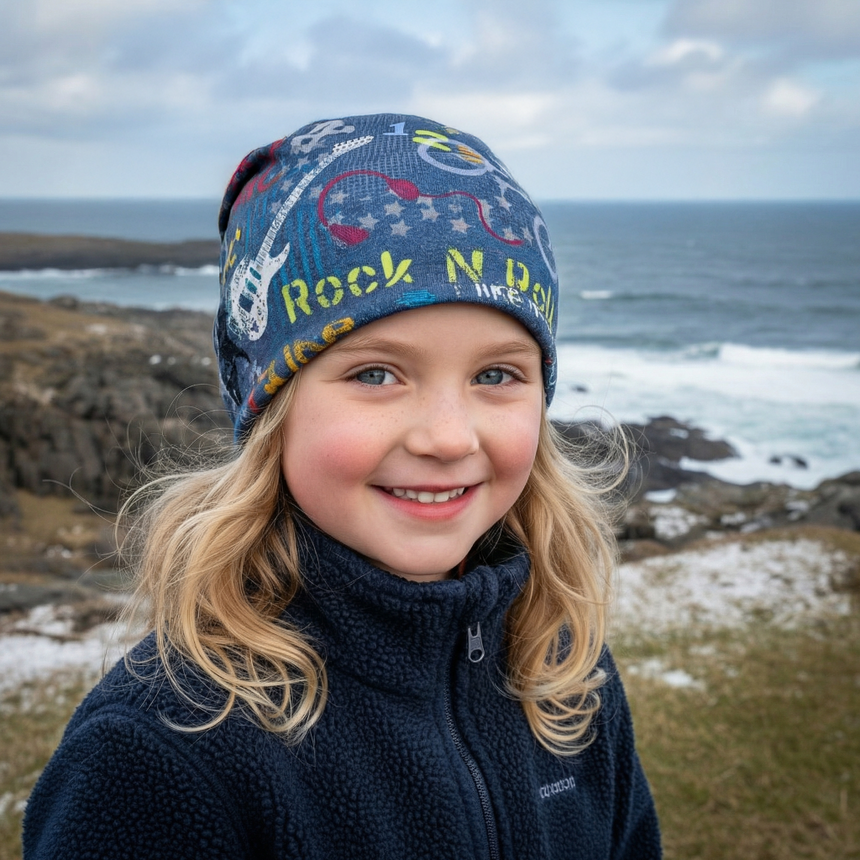 Mädchen mit Wollhuhn Öko Shorties Wendemütze am felsigen Küstenrand, lächelt in die Kamera. Beanie mit Gitarrenmotiv und Rock N Roll Aufschrift, handgemacht in Deutschland.