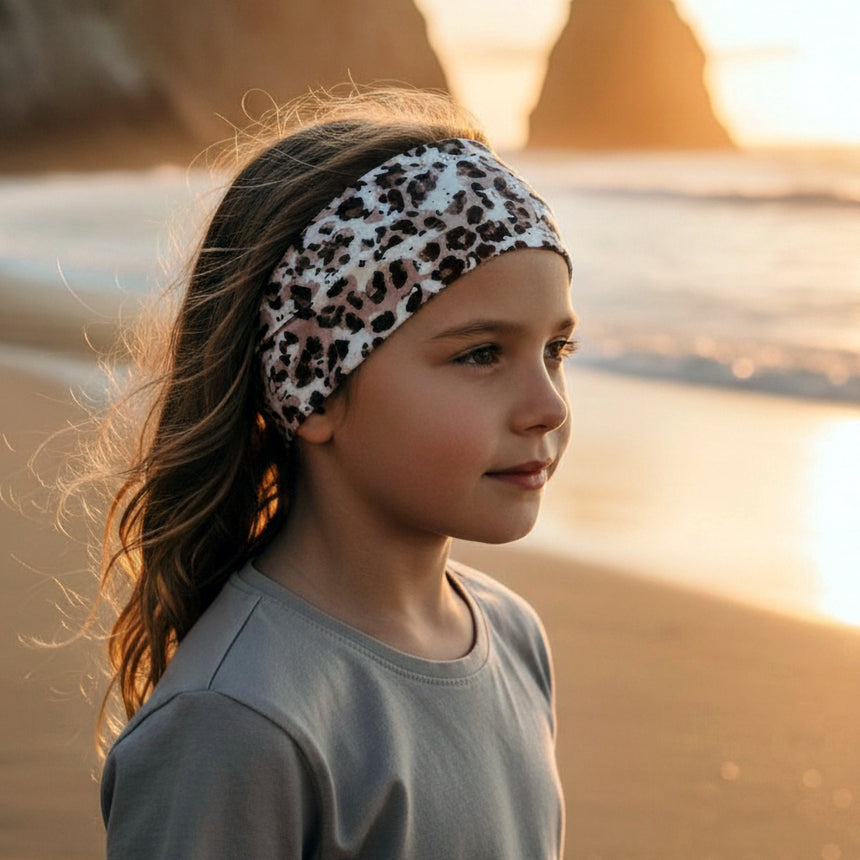 Mädchen am Strand trägt ein Wollhuhn Elastisches Öko Stirnband in Leopardendruck. Das handgemachte, breite Jersey-Stirnband aus weicher Bio-Baumwolle hält ihr Haar zurück.