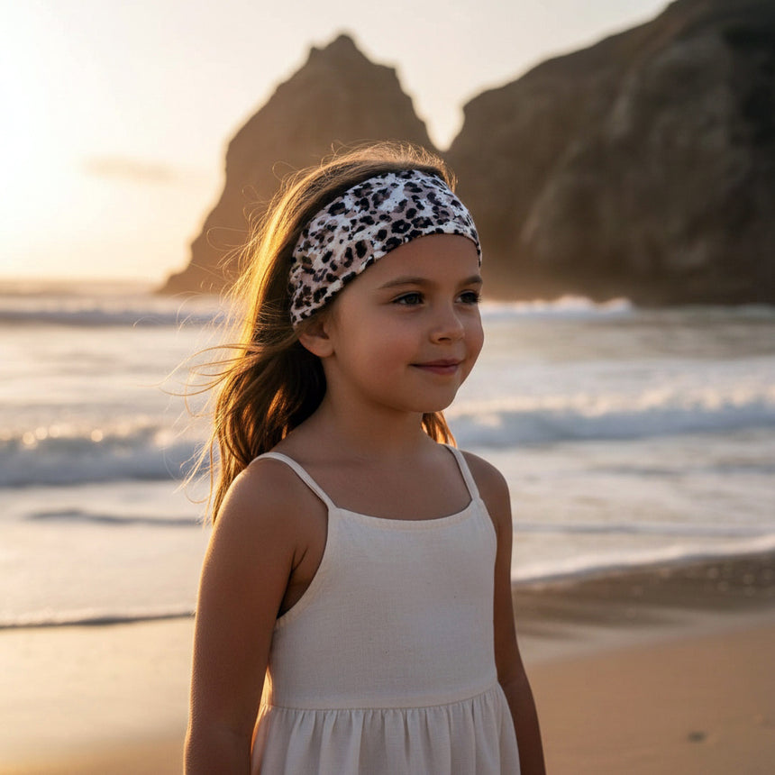 Mädchen am Strand bei Sonnenuntergang trägt Wollhuhn Elastisches Öko Stirnband, Jersey, zweilagig, breit, mit Leo-Muster – handgemacht und vielseitig tragbar.