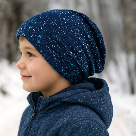 Kind mit Wollhuhn Öko Long Beanie, Galaxy Sprenkel Blau, aus zweilagigem Öko-Jersey, wendbar, ideal für milde Tage, vor verschneitem Waldhintergrund.