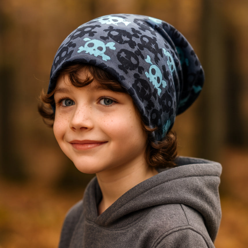 Kind mit lockigem Haar trägt Wollhuhn Öko Long Beanie, Wendemütze mit Totenkopf-Motiv. Perfekt für Herbst, zweilagig und bequem, hält warm. Ideal für Schule und Freizeit.