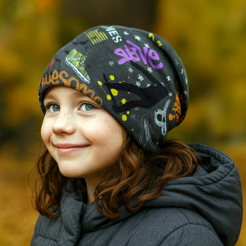 Ein Kind trägt die Wollhuhn Öko Long Beanie, dunkelgrau mit bunten Graffiti, lässig fallend, ideal für Herbsttage. Perfekt für Schule und Freizeit.