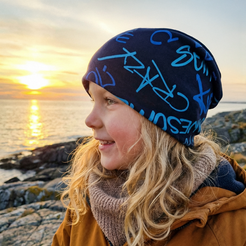 Kind am felsigen Ufer im Wollhuhn Öko Long Beanie, Cool Music Blautöne, blickt in den Sonnenuntergang. Bequem, wendbar, ideal für Übergangswetter.