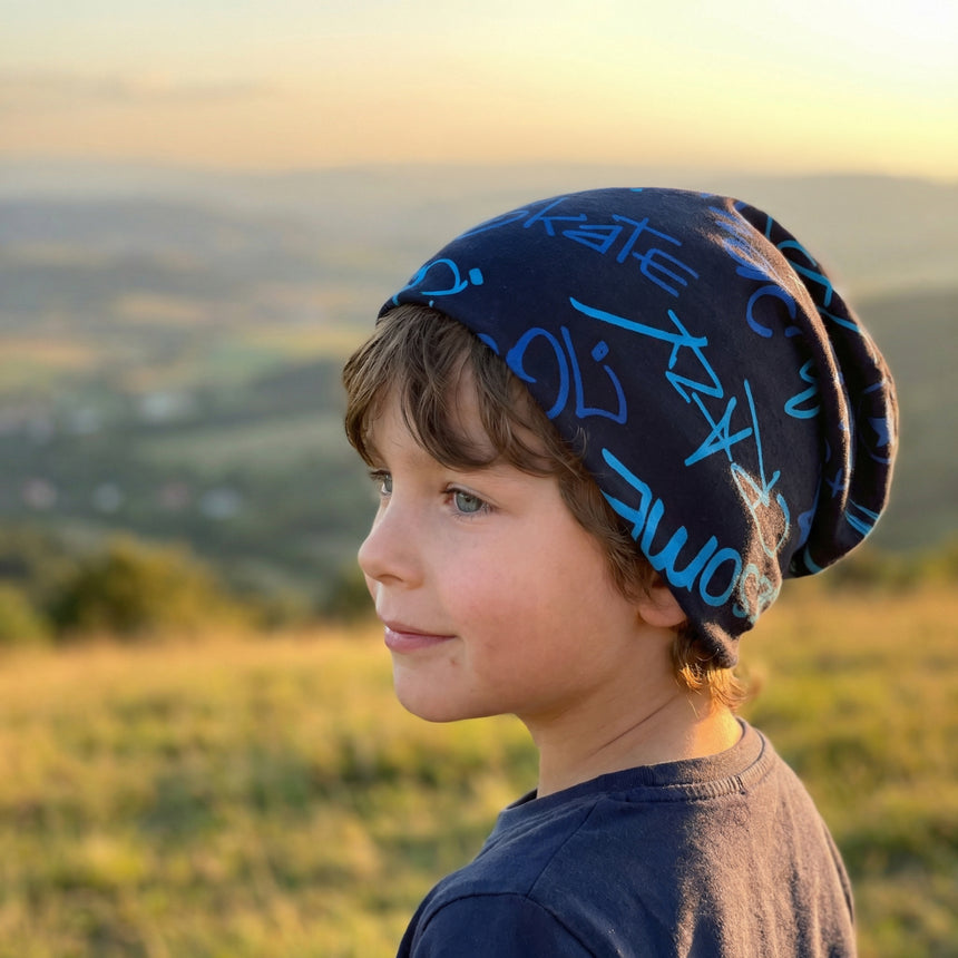 Junger Junge trägt Wollhuhn Öko Long Beanie mit blauer Graffiti-Schrift, lässig im Sonnenuntergang stehend, ideal für stilvolle, bequeme Outdoor-Aktivitäten.