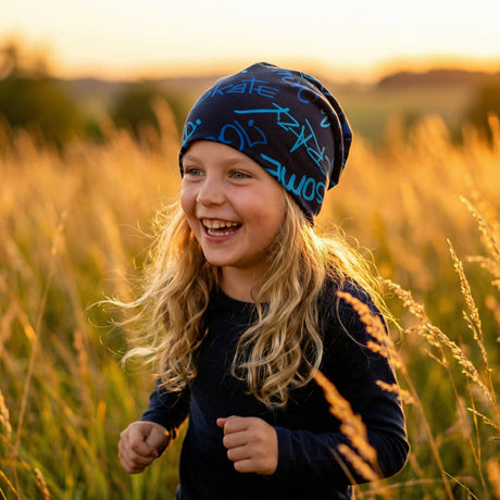 Kind mit blonden Haaren trägt eine Wollhuhn Öko Long Beanie / Wendemütze, Cool Music Blautöne, während es lachend durch ein sonnenbeschienenes Feld läuft.