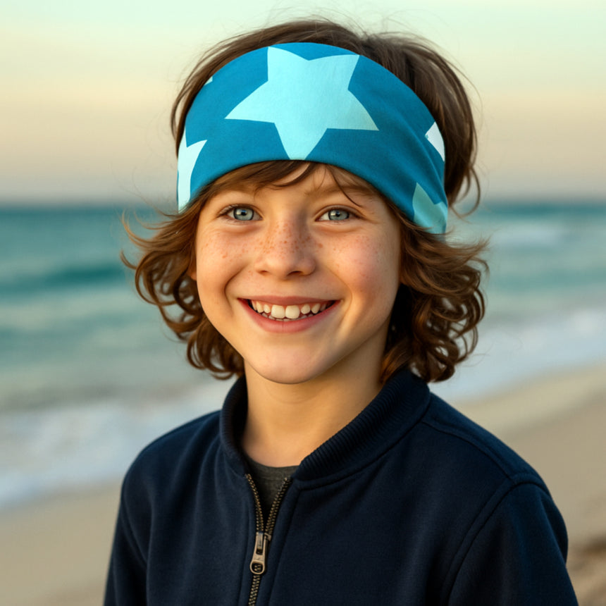 Kind am Strand trägt das Wollhuhn Elastisches Öko Stirnband/Haarband Jersey Zweilagig Breit Big Stars Sterne Petrol, ideal für vielseitige, nachhaltige Nutzung.