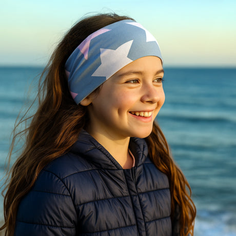 Mädchen am Meer trägt Wollhuhn Elastisches Öko Stirnband/Haarband Jersey Zweilagig Breit Big Stars Grau/Rosa, lächelnd bei Sonnenaufgang.