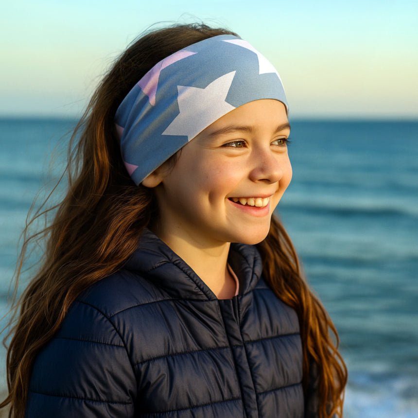 Mädchen am Meer trägt Wollhuhn Elastisches Öko Stirnband/Haarband Jersey Zweilagig Breit Big Stars Grau/Rosa, lächelnd bei Sonnenaufgang.