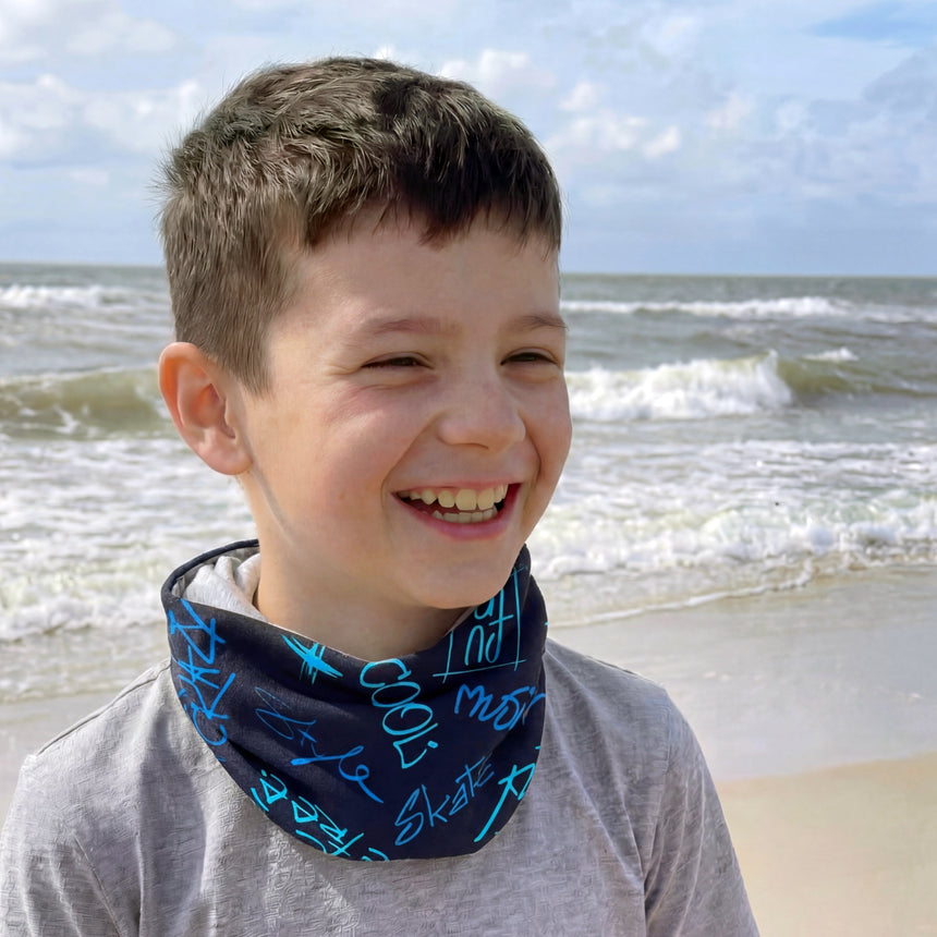Junger Junge am Strand trägt Wollhuhn Leichtes Lockeres Öko Jersey Halstuch/Schlupfschal Cool Music Blau, lächelnd, mit Wellen im Hintergrund. Perfekt für Übergangszeiten.