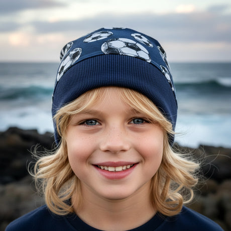 Kind mit blonden Haaren trägt Wollhuhn Dünne Öko Beanie-Mütze Sommer/Übergang Einlagig Fußball Dunkelblau, lächelt am Meer. Ideal für Übergangszeit, handgemacht in Deutschland.