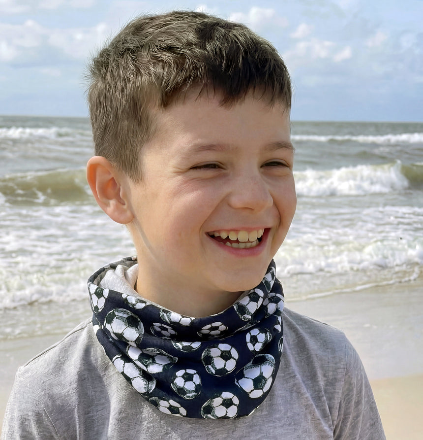 Junge am Strand trägt Wollhuhn leichtes Jersey Halstuch mit Fußballmuster, perfekt für Übergangszeiten, handgemacht in Deutschland, schützt vor Wind und Sonne.