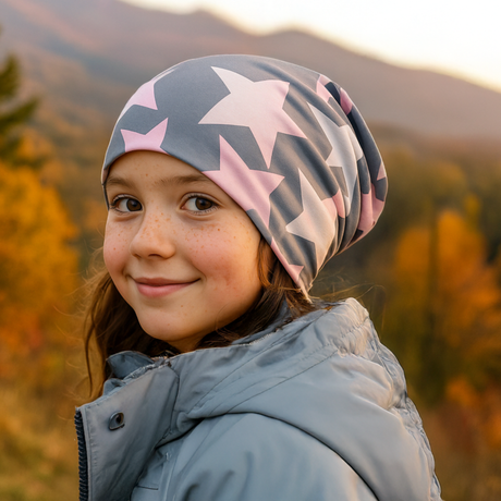Mädchen mit Wollhuhn Öko Long Beanie, Wendemütze, zweilagig, Big Stars Grau/Rosa/Pink, lächelt in herbstlicher Landschaft. Trägt graue Pufferjacke und Beanie mit Sternmuster.