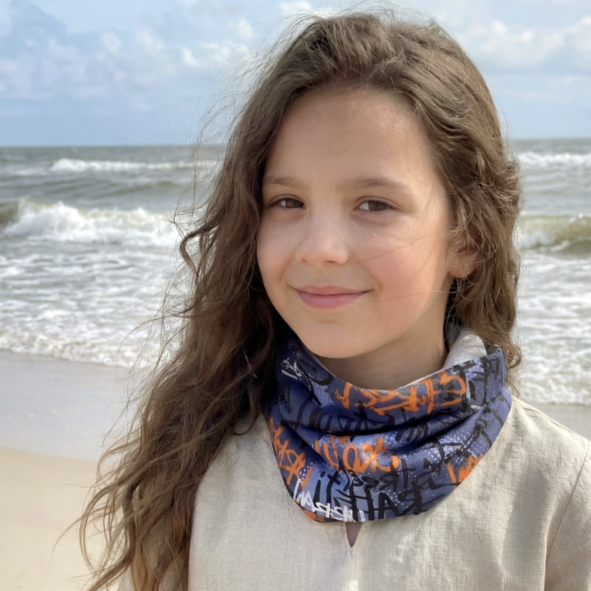 Mädchen am Strand trägt Wollhuhn Leichtes Lockeres Öko Jersey Halstuch in Grau/Orange, ideal für Übergangszeiten. Handgemacht in Deutschland, schützt vor Wind und Sonne.