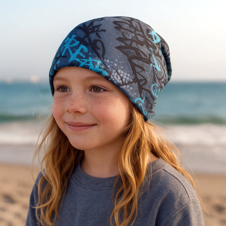 Mädchen am Strand trägt Wollhuhn Öko Shorties, Wendemütze, zweilagig, ideal für Kinder, handgefertigt aus Öko-Jersey, bequem und stylisch.