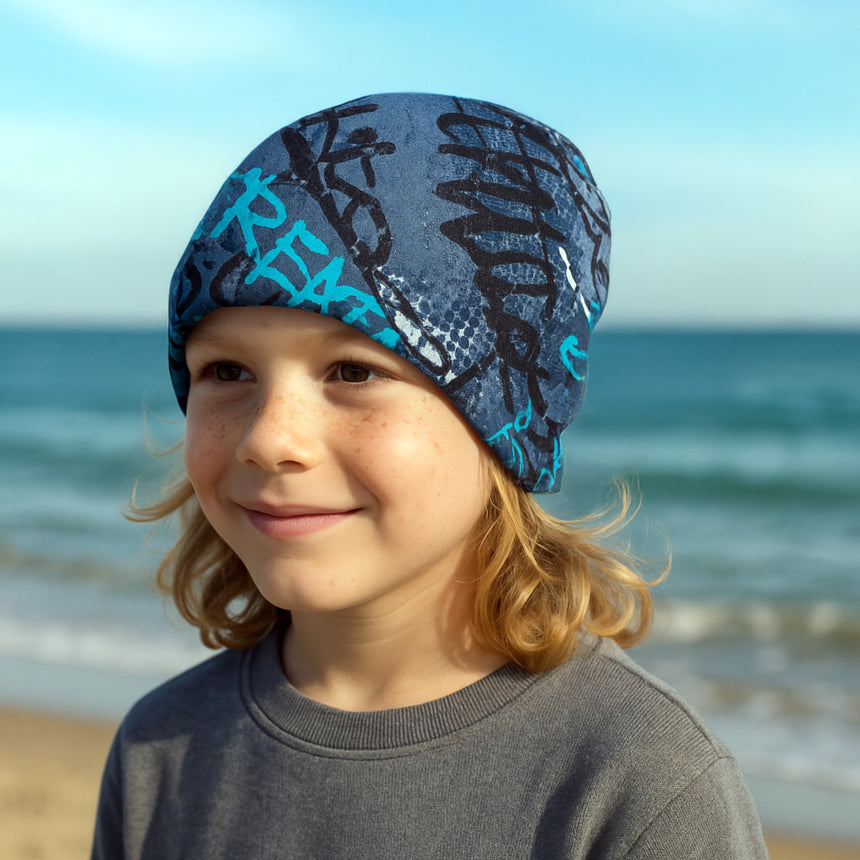 Kind am Strand mit Wollhuhn Öko Shorties, einer handgemachten, zweilagigen Wendemütze in Skater Blau, ideal für aktive Kinder, stilvoll und bequem.