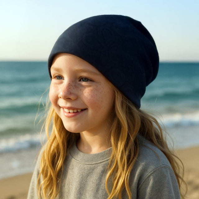 Mädchen am Strand trägt Wollhuhn Öko Shorties, eine handgemachte, zweilagige, wendbare Mütze, ideal für aktive Kinder, gefertigt aus Öko-Jersey.