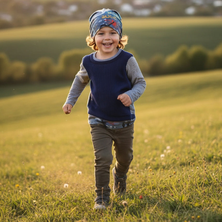 Ein Kleinkind trägt eine Wollhuhn Öko Long Beanie, Wendemütze, beim Laufen durch eine Wiese, lächelnd und gekleidet in bequemer Freizeitkleidung.