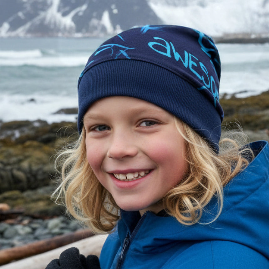 Kind am Felsenstrand, trägt Wollhuhn Dünne Öko Beanie-Mütze Sommer/Übergang Einlagig Cool Music Blau, lächelnd in blauer Jacke und awesome Beanie, handgemacht in Deutschland.