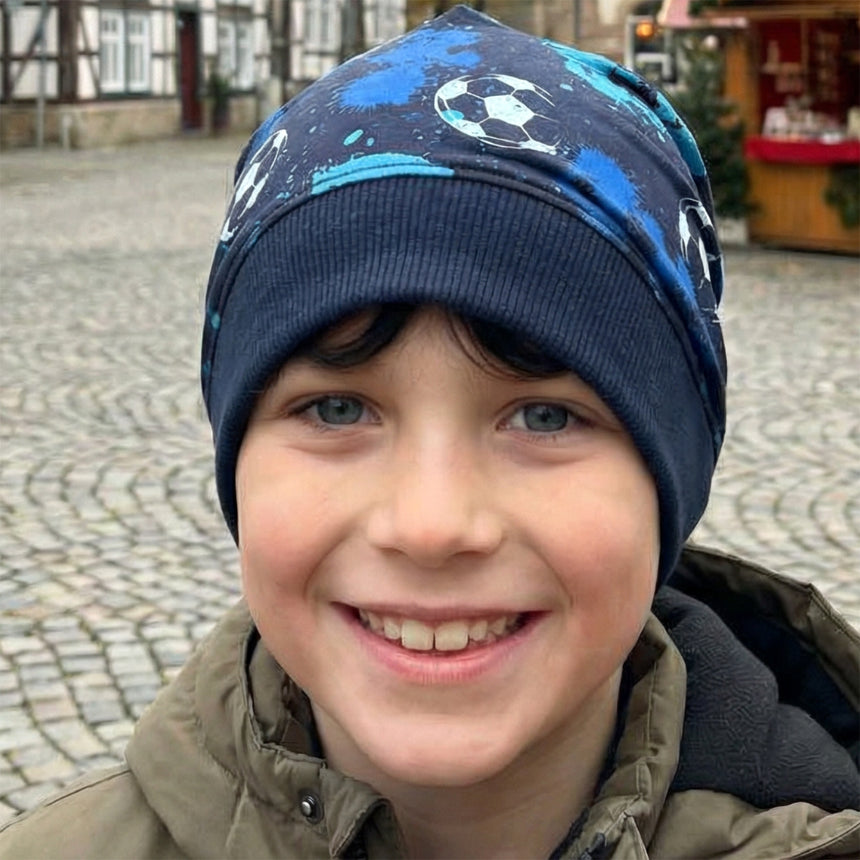 Junger Junge trägt Wollhuhn Dünne Öko Beanie-Mütze mit Fußballmotiv. Handgemacht aus Öko-Jersey, ideal für Übergangszeit, flexibel und bequem.