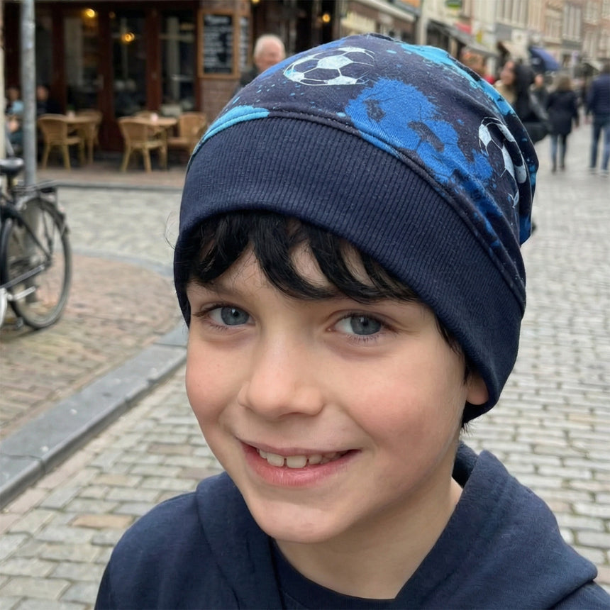 Junger Junge trägt Wollhuhn Dünne Öko Beanie-Mütze, Fußballmotiv in Blautönen, auf einer belebten Straße. Perfekt für Übergangszeiten, handgemacht aus Öko-Jersey.