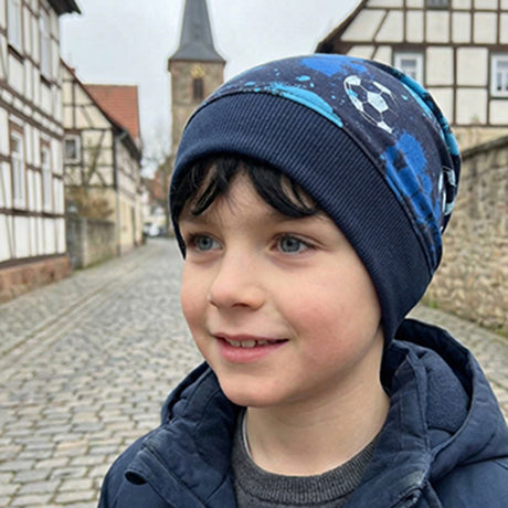 Junge trägt Wollhuhn Dünne Öko Beanie-Mütze Sommer/Übergang Einlagig Fußball Blautöne auf Kopfsteinpflasterstraße, umgeben von Fachwerkhäusern und Kirchturm im Hintergrund.