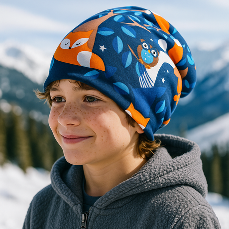 Ein Kind trägt die Wollhuhn Öko Long Beanie / Wendemütze, Nightfox Füchse Dunkelblau/Orange, in einer verschneiten Berglandschaft, passend für milde Wintertage.