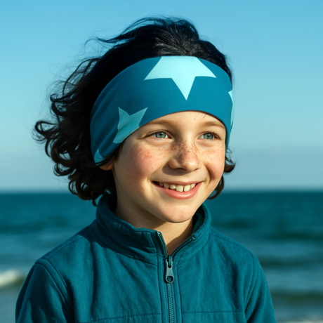 Ein freudiges Kind am Meer trägt das Wollhuhn Elastisches Öko Stirnband/Haarband Jersey Zweilagig Breit Big Stars Sterne Petrol, ideal für aktive Outdoor-Abenteuer.
