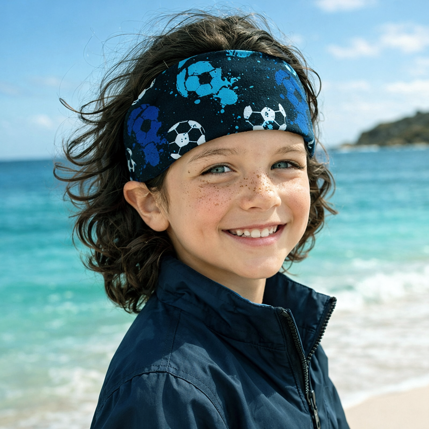Ein Kind am Strand trägt das Wollhuhn Elastisches Öko Stirnband, zweilagig und breit, mit blauem Fußball-Design, perfekt für windige Outdoor-Aktivitäten.