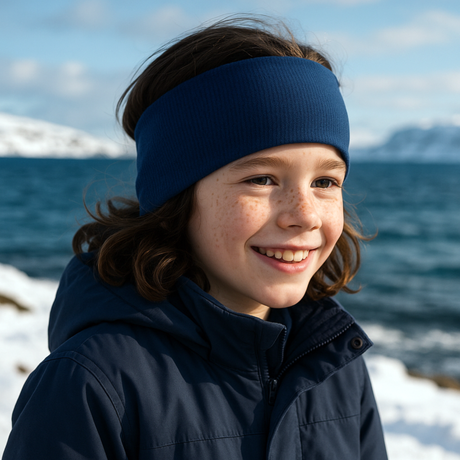 Kind trägt Wollhuhn Stirnband aus royalblauem Sportfleece, lächelnd in winterlicher Küstenlandschaft. Stirnband handgefertigt in Deutschland, zweilagig und windabweisend.