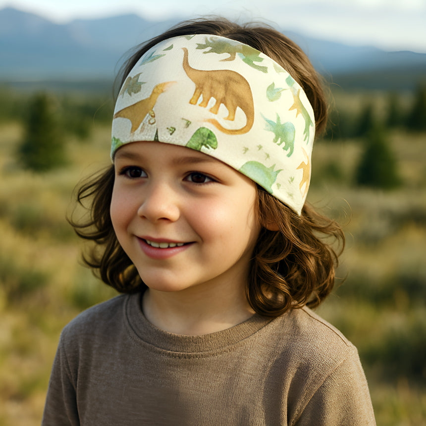 Kind trägt Wollhuhn Elastisches Öko Stirnband, Haarband Jersey Zweilagig Breit mit Dinosaurier-Print. Ideal für Outdoor-Aktivitäten, handgemacht aus Bio-Baumwolle, weich und dehnbar.