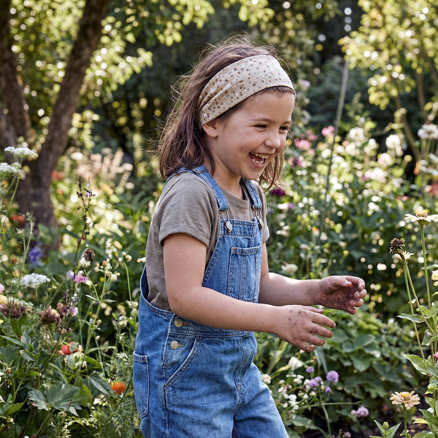 Mädchen in Garten trägt Wollhuhn Elastisches Öko Stirnband Jersey Zweilagig Breit Blümchen Sand, lächelt, umgeben von Blumen, zeigt Freude an Outdoor-Aktivitäten und stilvollem Komfort.