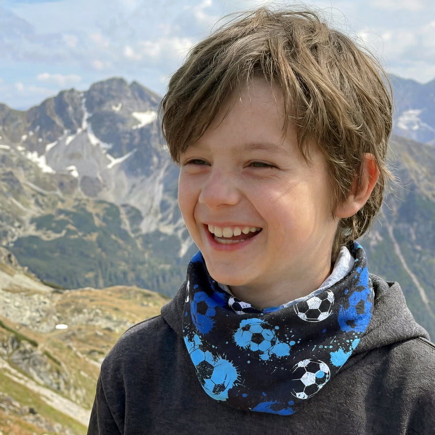 Ein lächelnder Junge trägt das Wollhuhn Leichtes/Lockeres Öko Jersey Halstuch, bedruckt mit Fußballmotiven, in einer bergigen Landschaft. Ideal für Übergangszeiten, handgemacht in Deutschland.