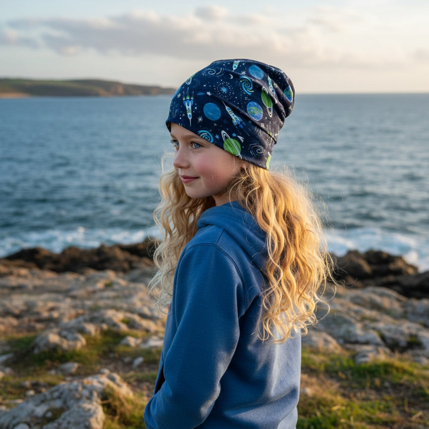 Mädchen am Meer mit Wollhuhn Öko Long Beanie, zweilagig, Space/Rakete-Muster, perfekt für Übergangszeiten. Handgefertigt, bequem und nachhaltig.