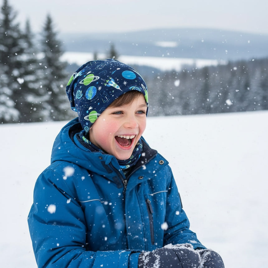 Junge mit Wollhuhn Öko Long Beanie, zweilagig, Space/Rakete Dunkelblau, lacht im Schnee, trägt blauen Wintermantel und hält Schnee in den Händen.