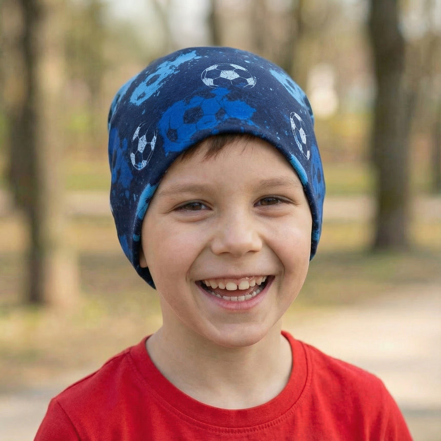 Junge mit Wollhuhn Öko Long Beanie, zweilagig, Fußballmuster in Blautönen, lächelt draußen. Mütze aus weichem Baumwolljersey, ideal für ganzjähriges Tragen.