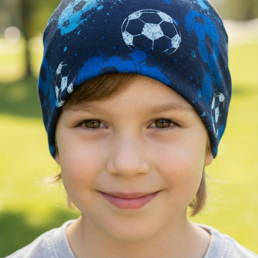 Kind mit Wollhuhn Öko Shorties Wendemütze, Fußball Blau, lächelt draußen. Beanie mit Fußballmuster, ideal für aktive Kinder. Handgemacht, zweilagig und komfortabel.